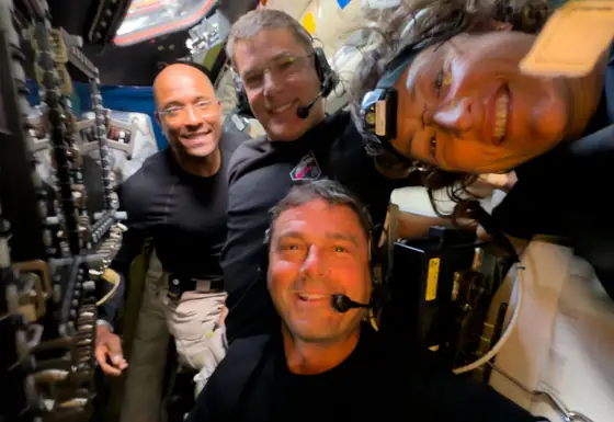 Quatre astronautes souriants, serrés tête contre tête entre les parois couvertes d’équipements de la capsule.