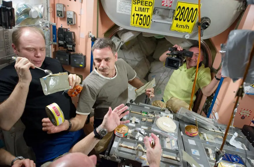 Trois cosmonautes partagent un repas en sachets autour d’une table, à bord de la Station spatiale internationale.